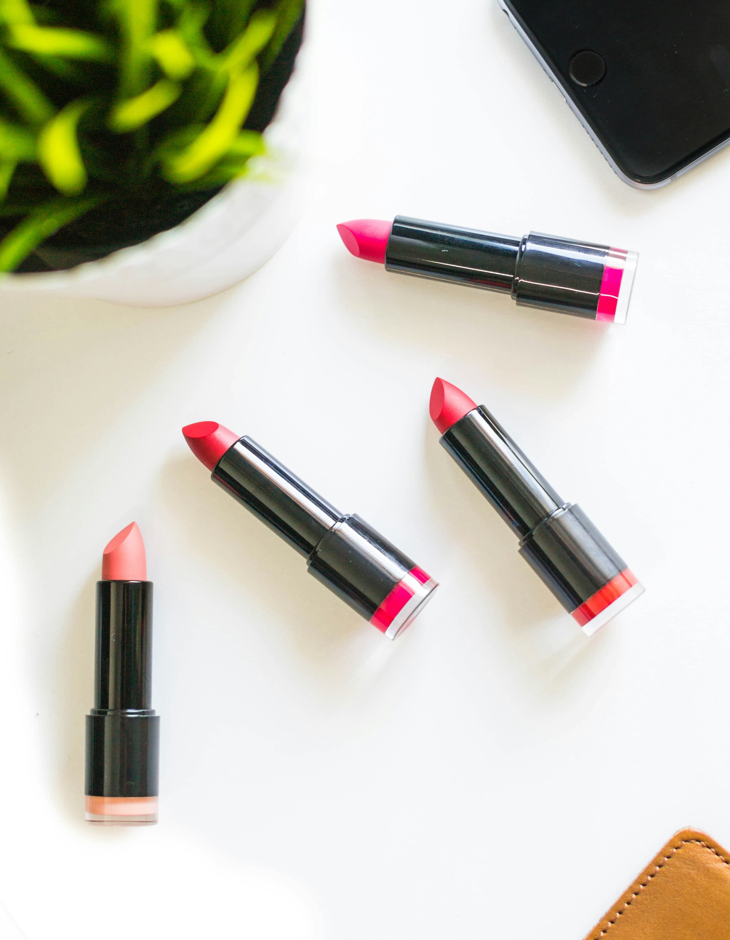 Four lipsticks in black cases, in nude, red and pink shades, on a white table with a plant and phone corner.