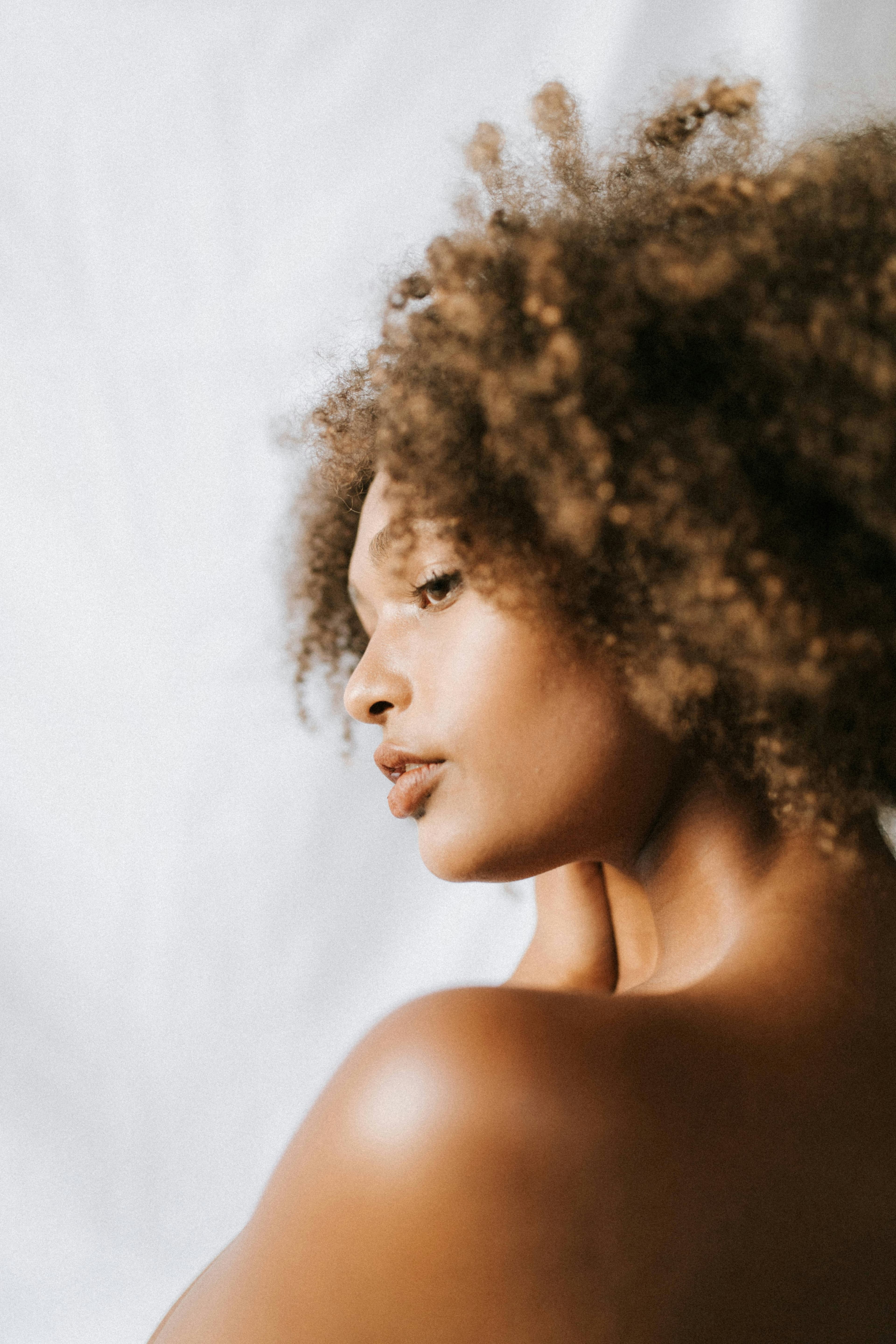 Profile of a woman with voluminous curly brown hair and bare shoulders against a soft white backdrop, warm daylight on her skin.