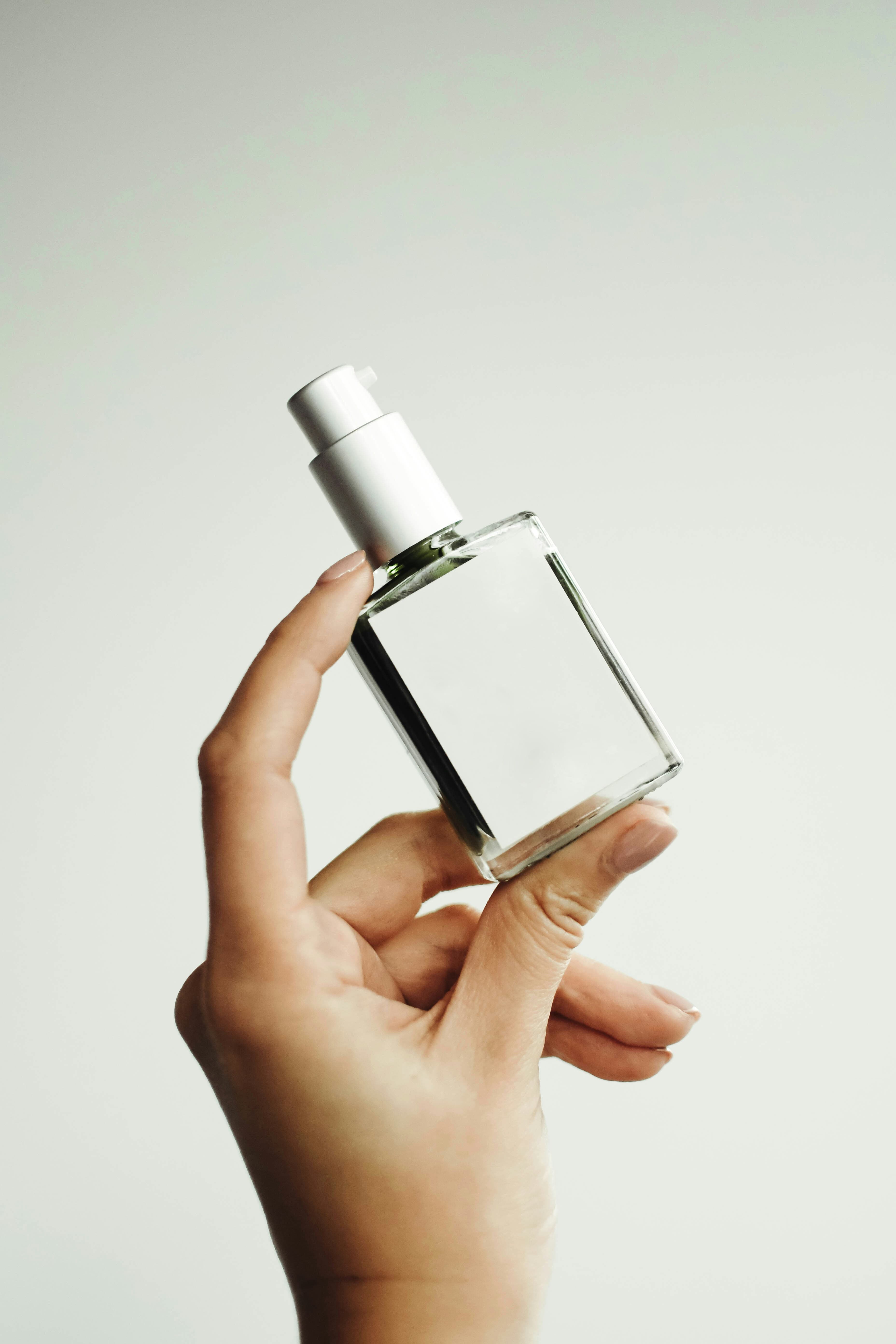 Hand holding a blank clear rectangular bottle with a white pump against a pale grey-green background.
