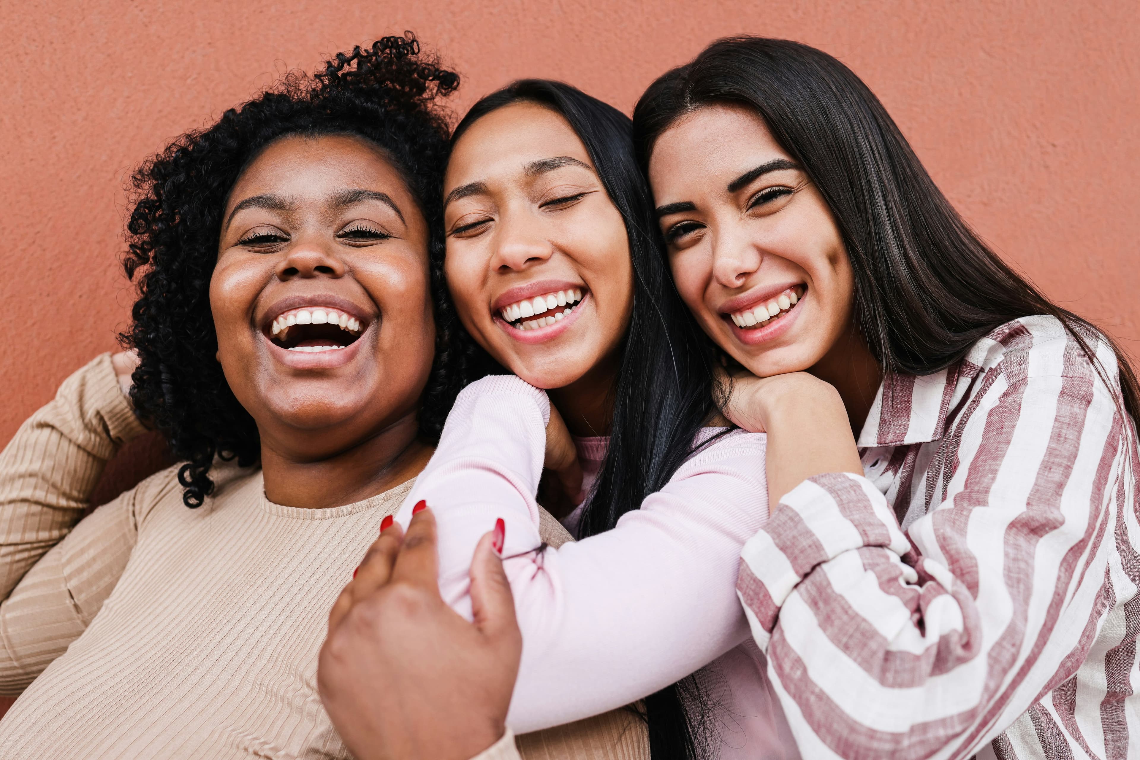 Three smiling women with different skin tones hugging and laughing together in front of a terracotta wall.