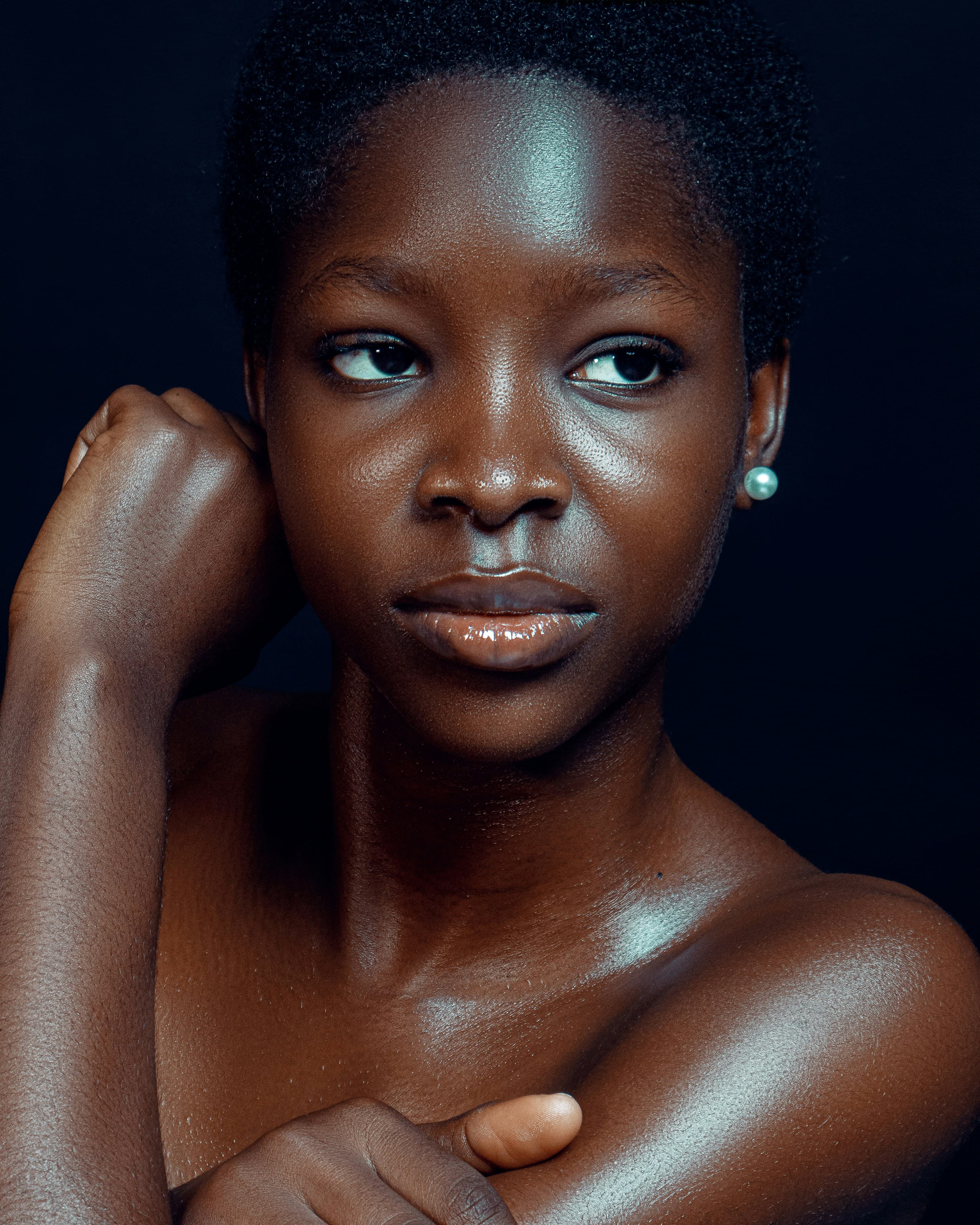 Portrait of a Black woman with close-cropped hair and glowing, dewy skin, hand raised near her face, against a deep navy backdrop.
